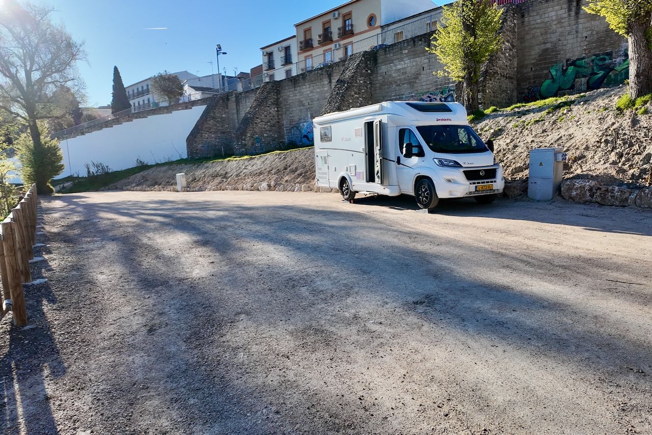 Een witte camper staat geparkeerd op een grindterrein bij een stenen muur, met op de achtergrond gebouwen zichtbaar op een heuvel.