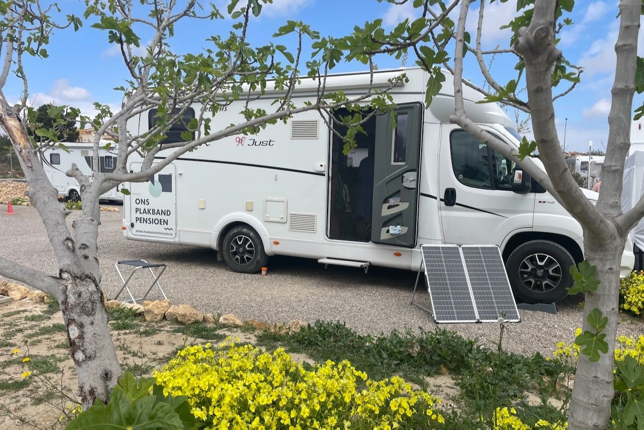 Een witte camper staat geparkeerd op een grindterrein vlakbij gele wilde bloemen, met verplaatsbare zonnepanelen bij de ingang.