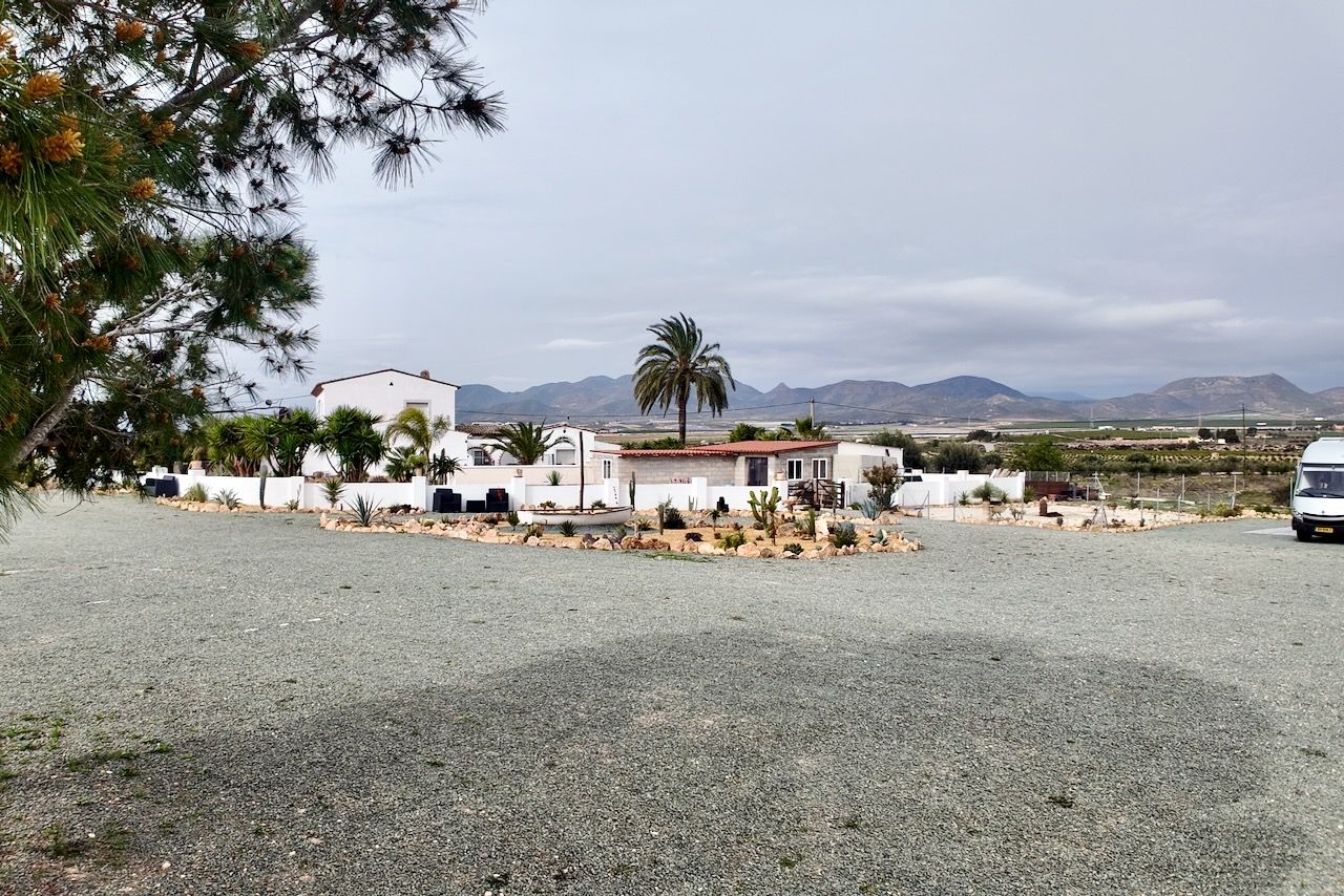 Een grindterrein met witte gebouwen, palmbomen en bergen onder een bewolkte hemel.