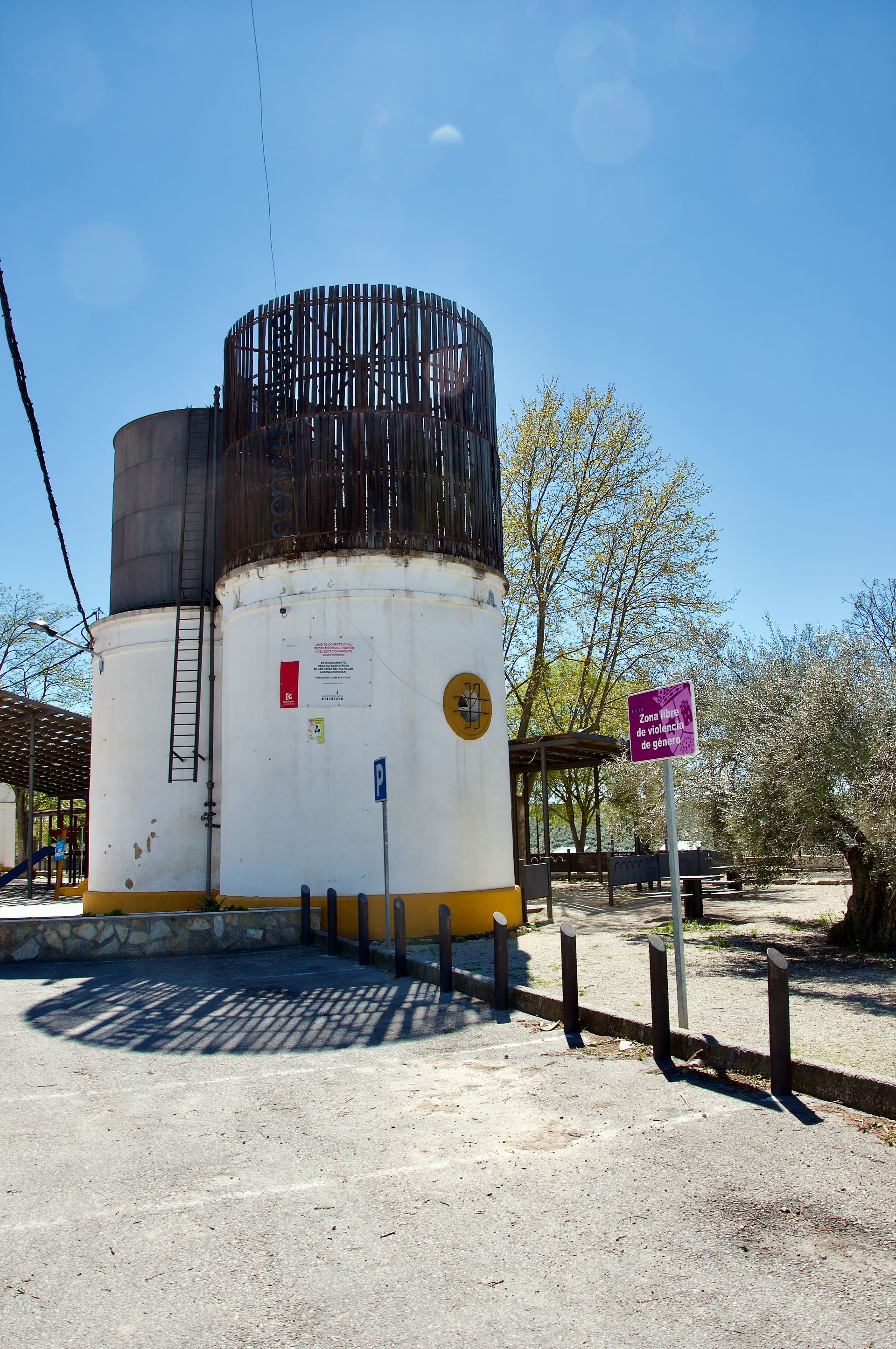 Een rustieke witte toren met een houten bovenbouw staat op een zonnige, met grind bedekte plek, vlakbij bomen en een klein bordje.