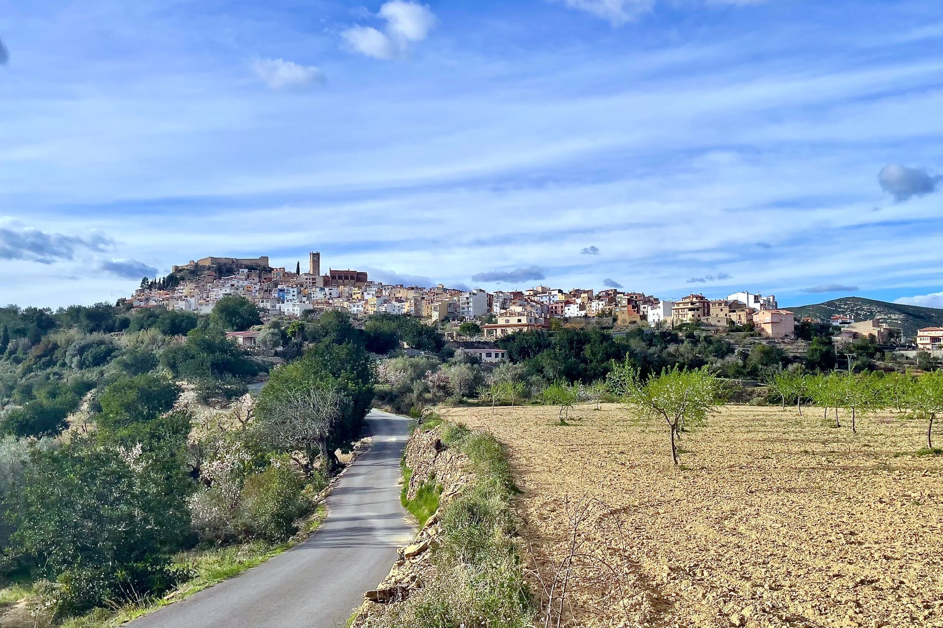 Een kronkelende weg leidt naar een dorpje op een heuveltop onder een bewolkte blauwe hemel. Bruine velden en groene bomen omringen het dorp.