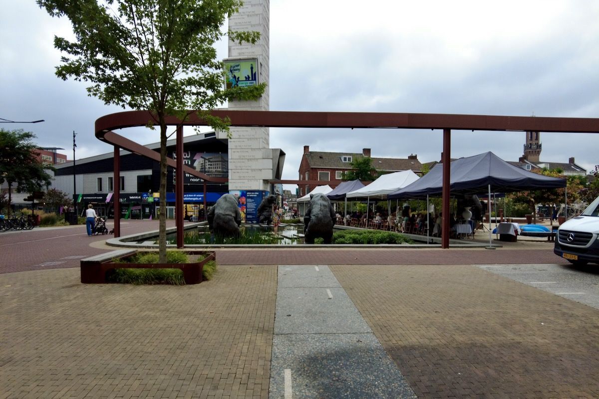 Buiten lunch Marktkraampjes en kunstsculpturen op een plein met een bruine stalen boog, onder een bewolkte hemel.