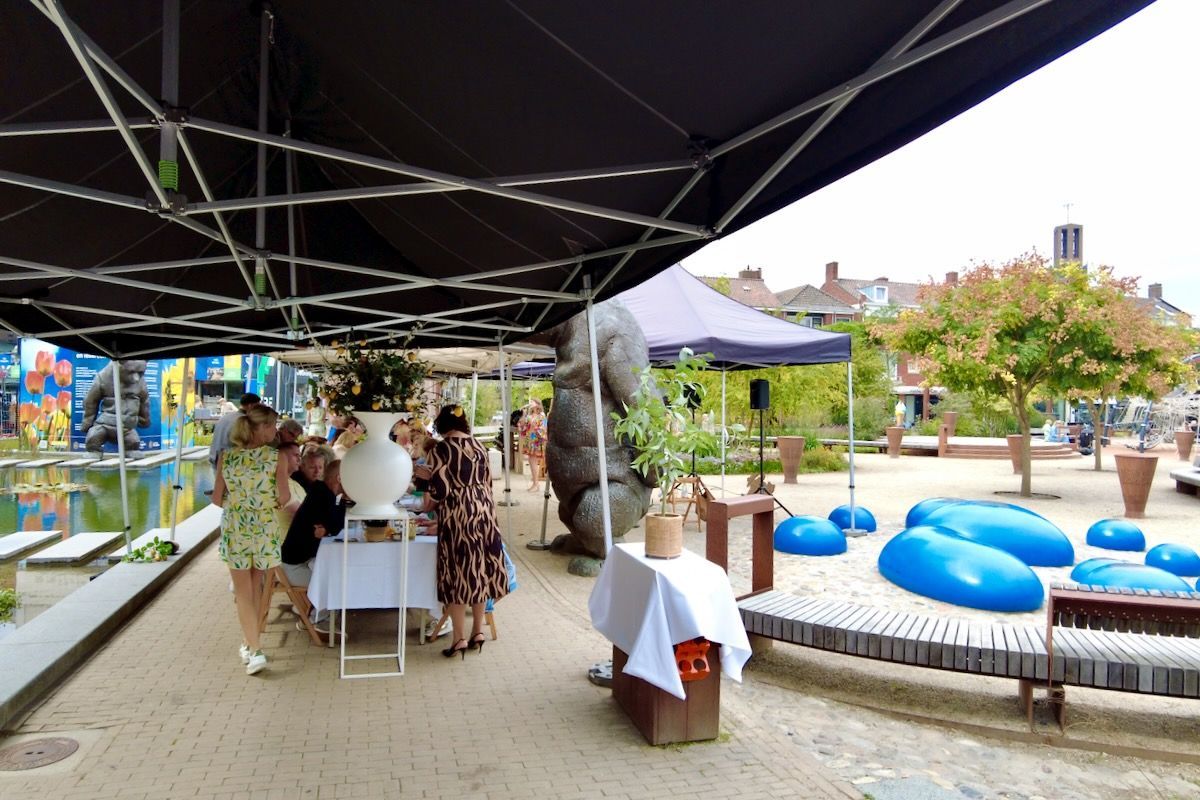 Gezellig hoor Mensen verzamelen zich voor een buitenevenement onder een zwart bladerdak. Er zijn blauwe sculpturen, water en een groot grijs gorillabeeld.