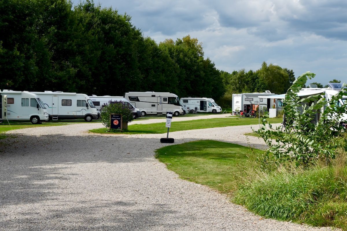 Overzicht Camperplaats met geparkeerde campers op grind, groen gras en bomen.