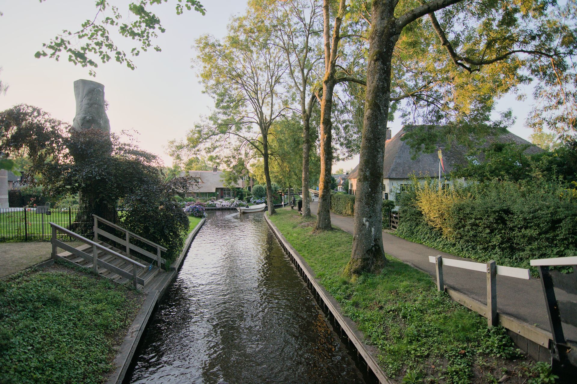 Een smal kanaal in Giethoorn, omzoomd door bomen en huizen, met een voetpad en een kleine brug bij zonsondergang.
