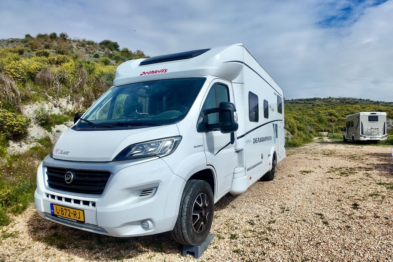 Een witte camper staat geparkeerd op een grindheuvel, met op de achtergrond een andere camper onder een blauwe hemel.