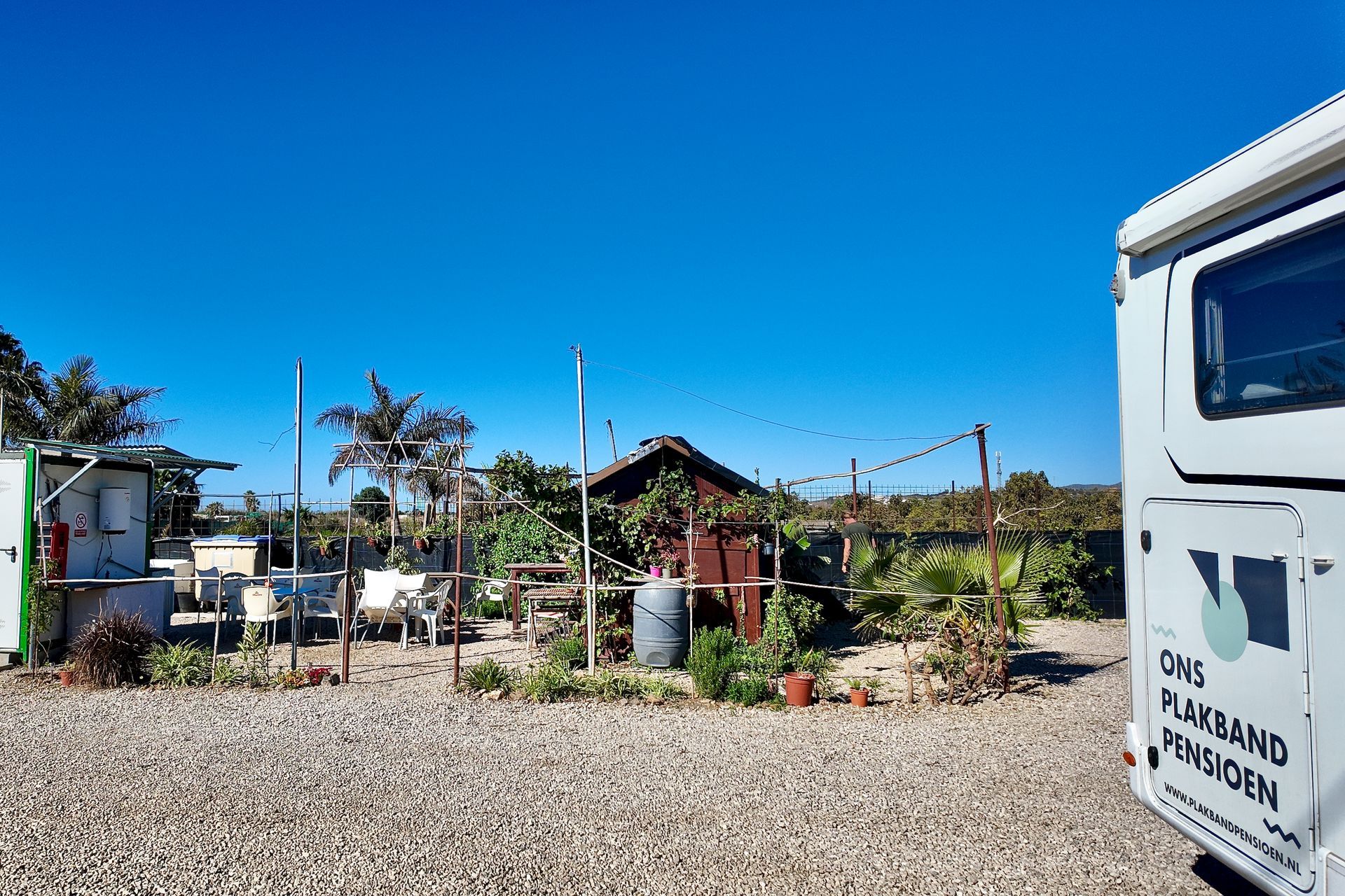 Een parkeerplaats met grind, een klein houten schuurtje, tuinmeubilair en een witte camper rechts geparkeerd onder een blauwe hemel.