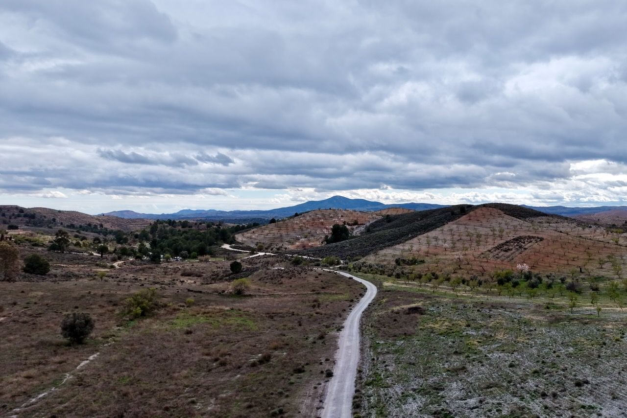Een zandpad leidt door een heuvelachtig, droog landschap naar verre bergen onder een bewolkte, grijze hemel.