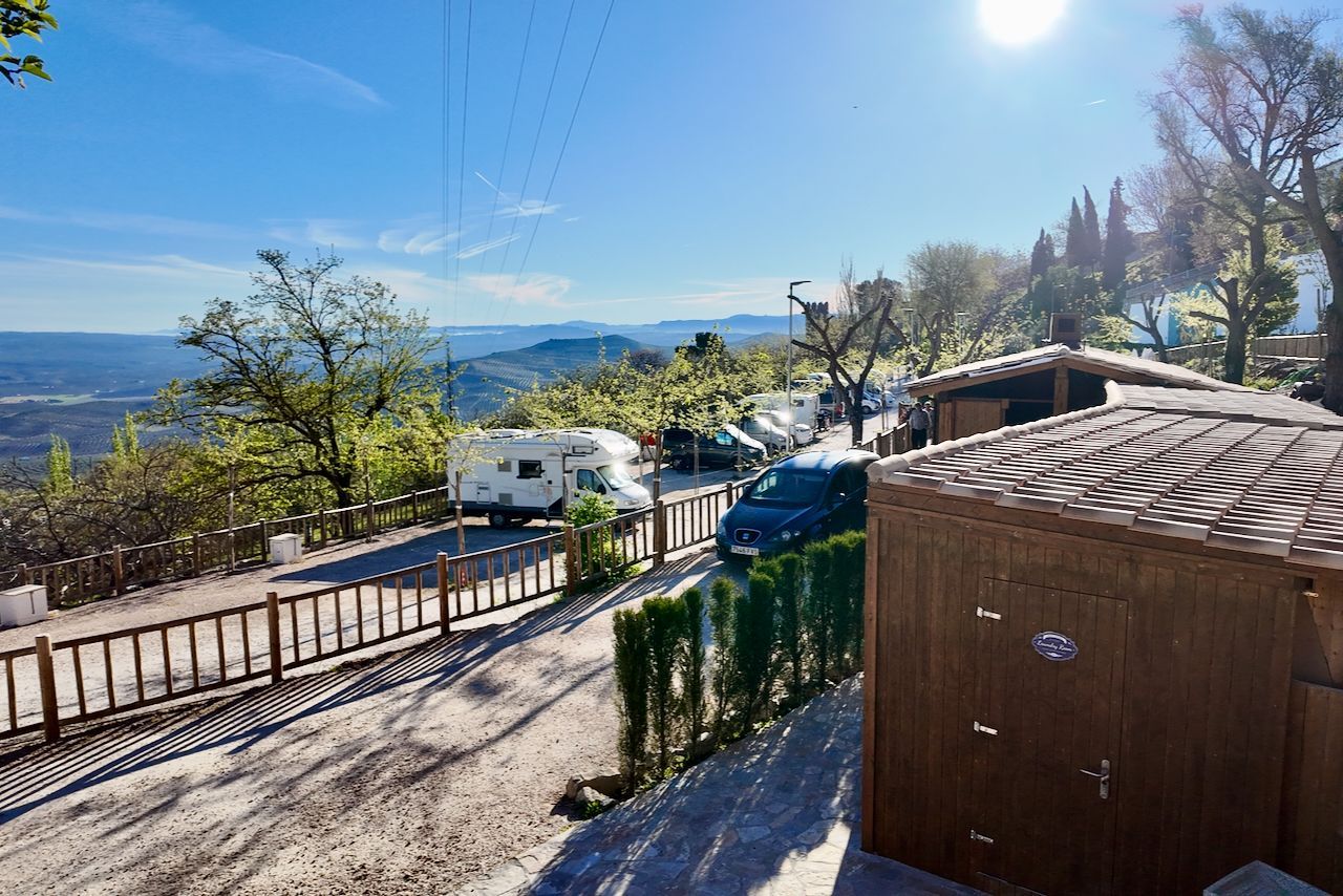 Een schilderachtig uitzicht op een zonnige camping op een heuvel met geparkeerde caravans, houten omheining en in de verte bergen onder een blauwe hemel.