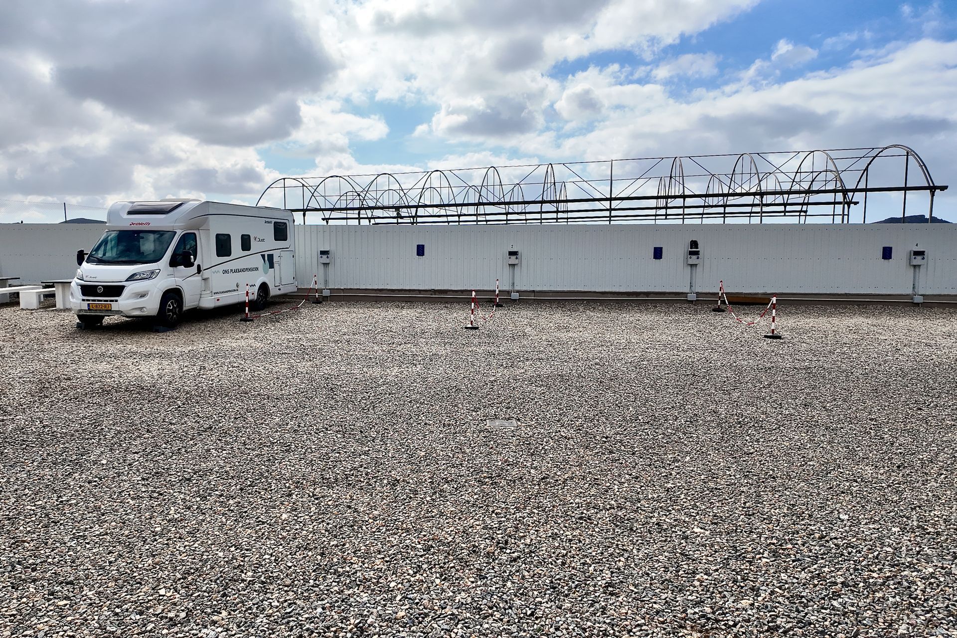 Een witte camper staat geparkeerd op een grindterrein voor een witte muur met daarop een draadhek, onder een bewolkte hemel.