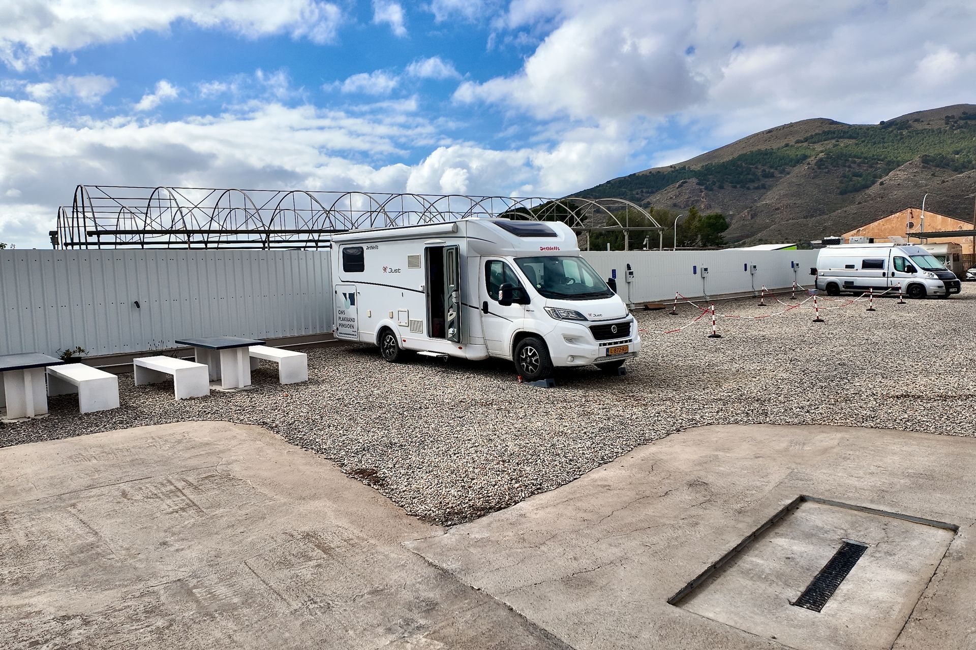 Een witte camper staat geparkeerd op een grindterrein naast een golfplaten hek, met een berg op de achtergrond.