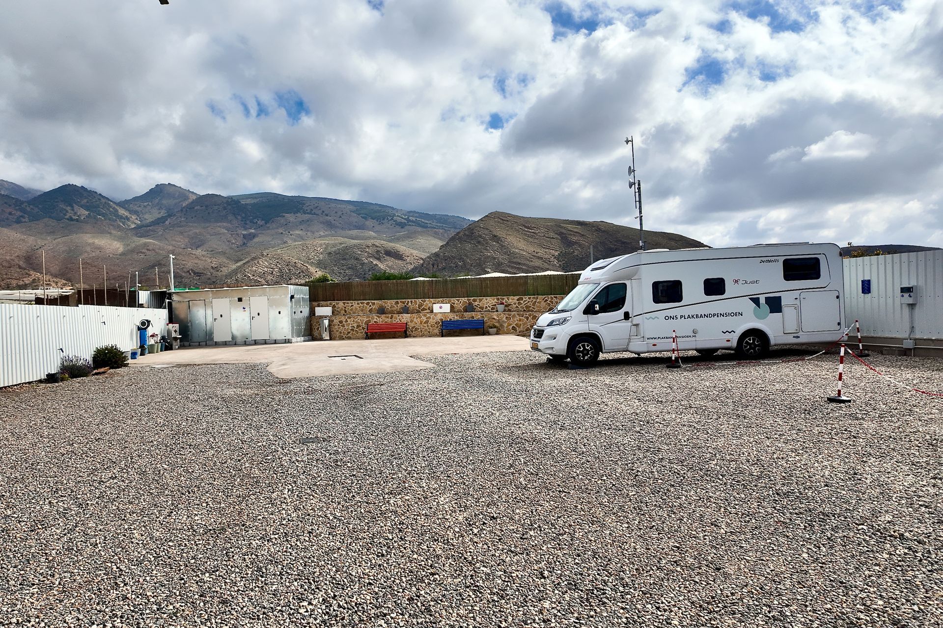 Een witte camper staat geparkeerd op een grindterrein onder een bewolkte hemel, met op de achtergrond dorre bergen.