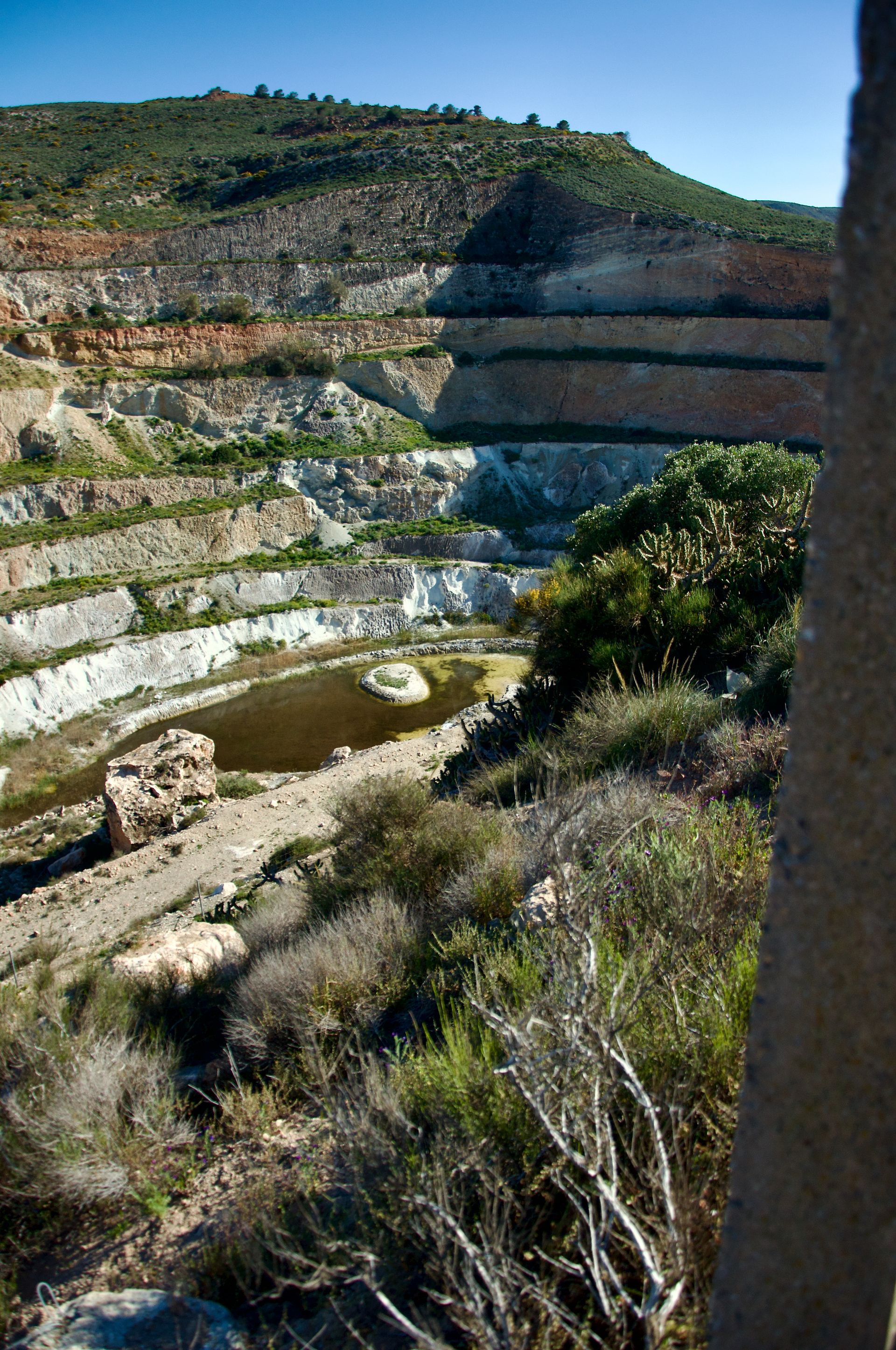 Een verlaten open mijn met terrasvormige rotswanden, schaarse vegetatie en een kleine waterpoel aan de voet.