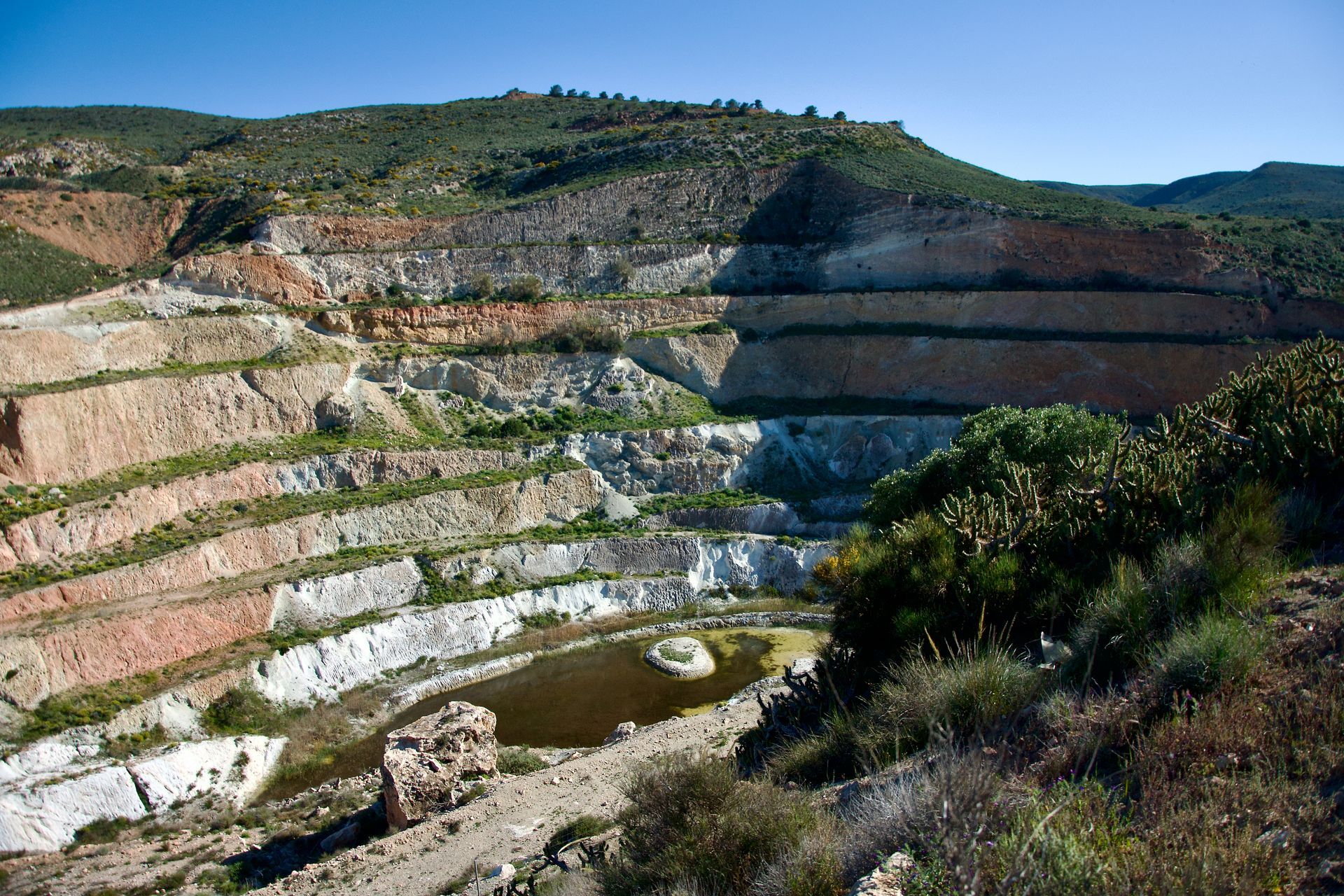 Een open mijn met terrasvormige, veelkleurige rotswanden en een kleine, troebele waterpoel aan de voet.
