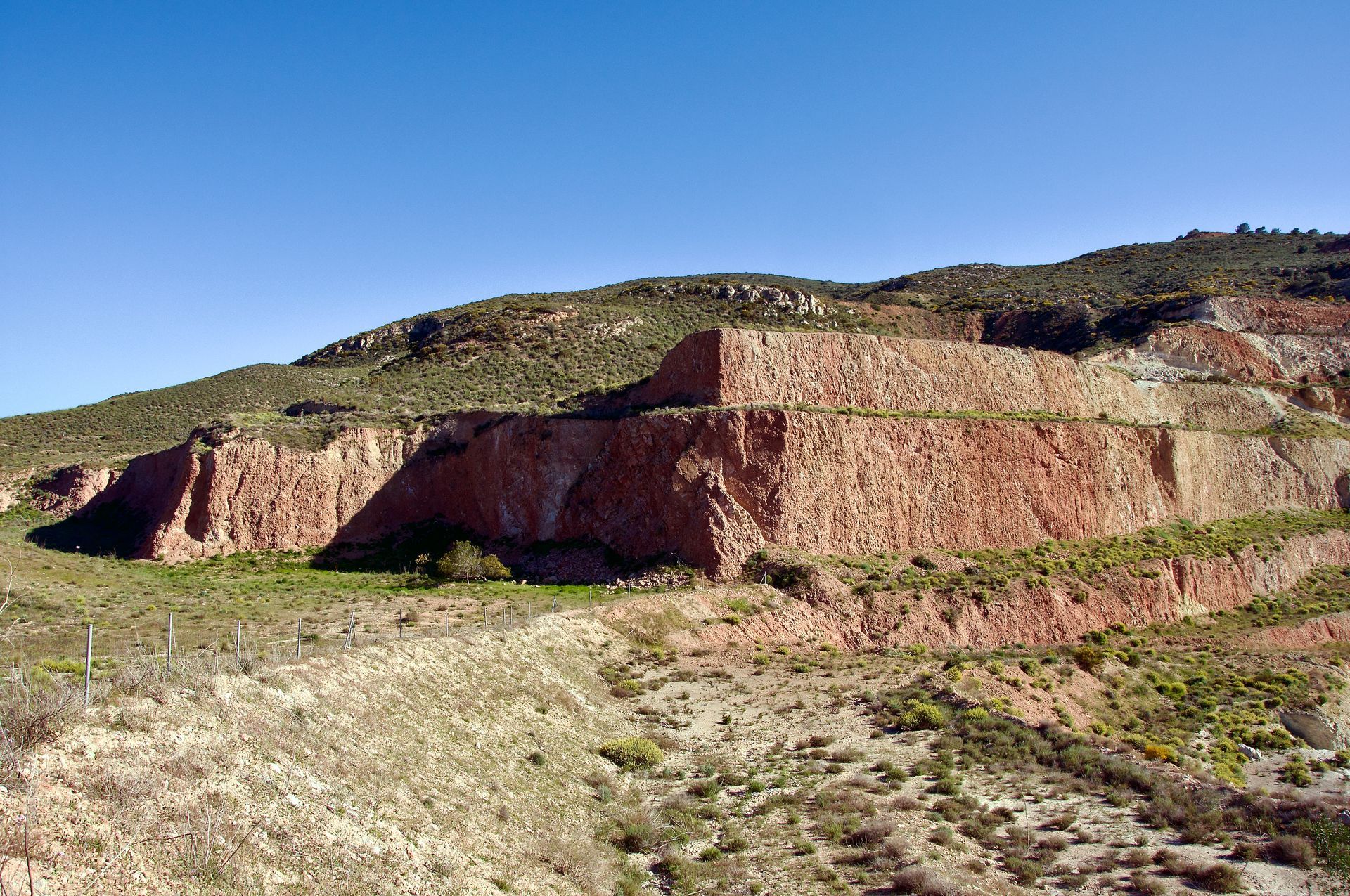 Een trapsgewijs aangelegde open mijn met blootliggende rode rotswanden tegen een helderblauwe hemel.