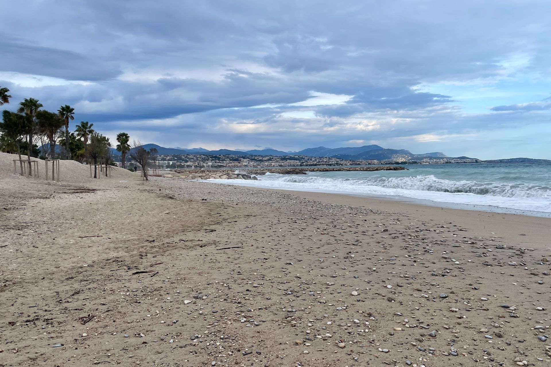 Het strand Cagnes sur Mer Een zandstrand met kleine golfjes onder een bewolkte hemel. Palmbomen en gebouwen in de verte zijn zichtbaar.