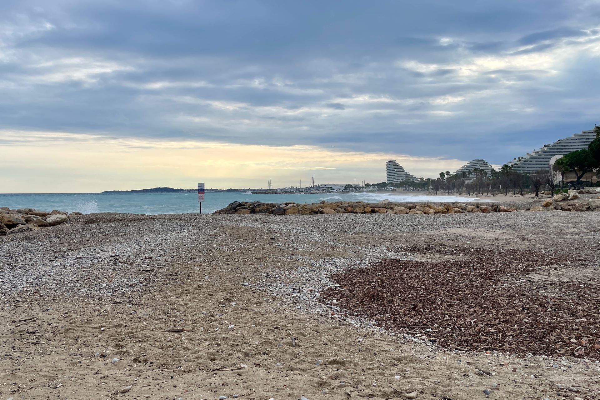 Het strand Villeneuve Loubet Strandtafereel met een bewolkte lucht, golven en een rotsachtige kust; gebouwen op de achtergrond.