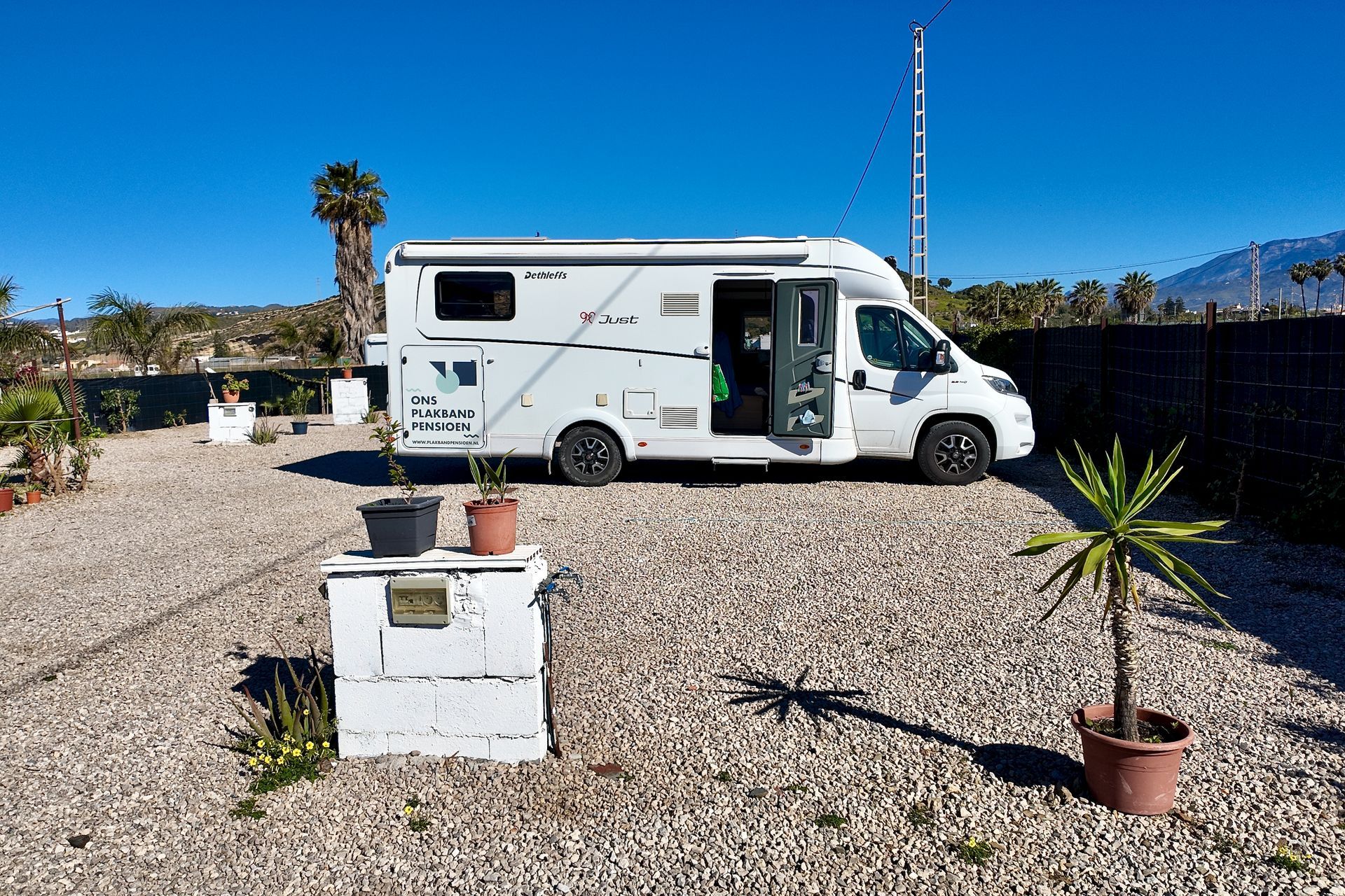 Een witte camper staat geparkeerd op een grindterrein onder een helderblauwe hemel, met een kleine palmboom in een pot en een elektriciteitsblok.