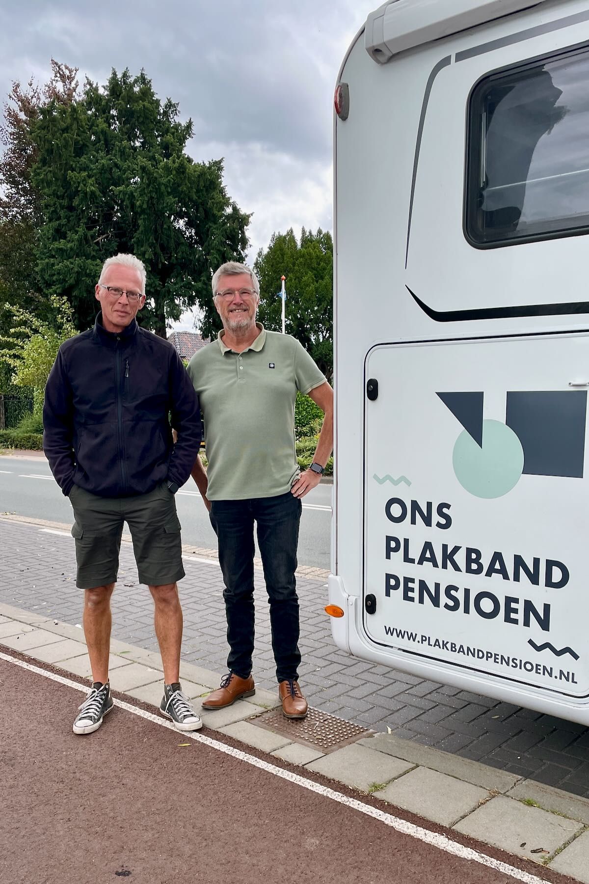 Gerhard Hormann Twee mannen staan naast een witte camper. De een draagt een korte broek, de ander een spijkerbroek. Op de camper staat: 'ONS PLAKBAND PENSIOEN'.