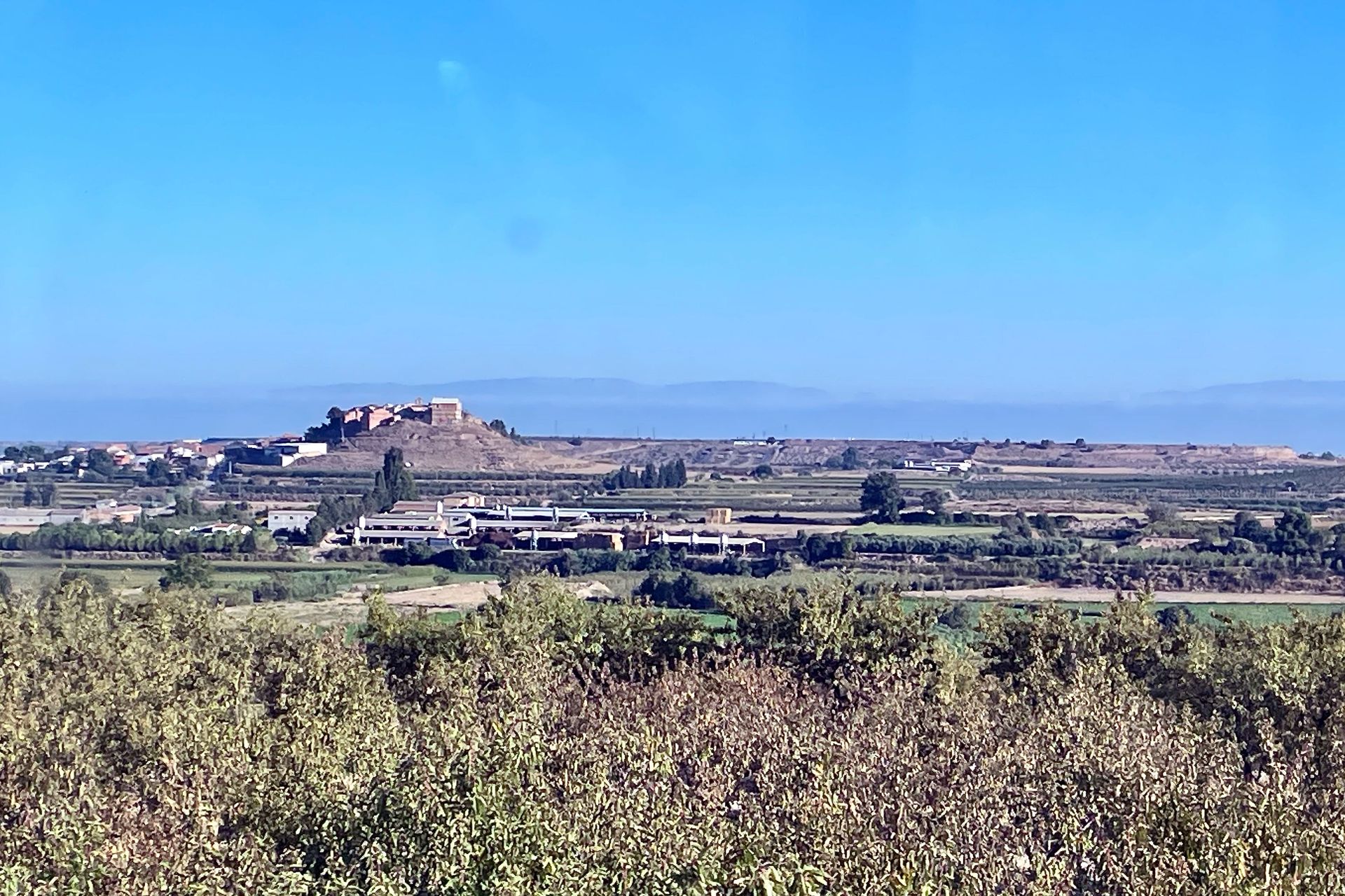 Vergezicht op een fort bovenop een heuvel, met gebouwen en velden op de voorgrond onder een helderblauwe lucht.