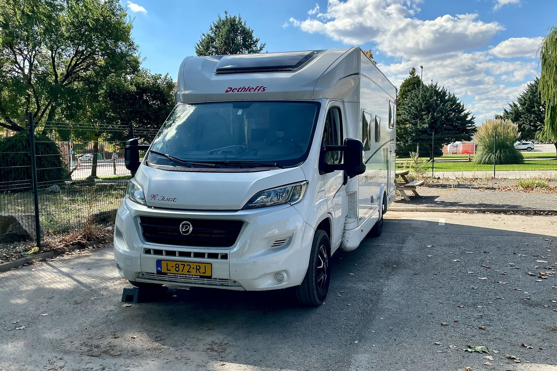 Witte camper geparkeerd op grind met bomen en hek op de achtergrond.