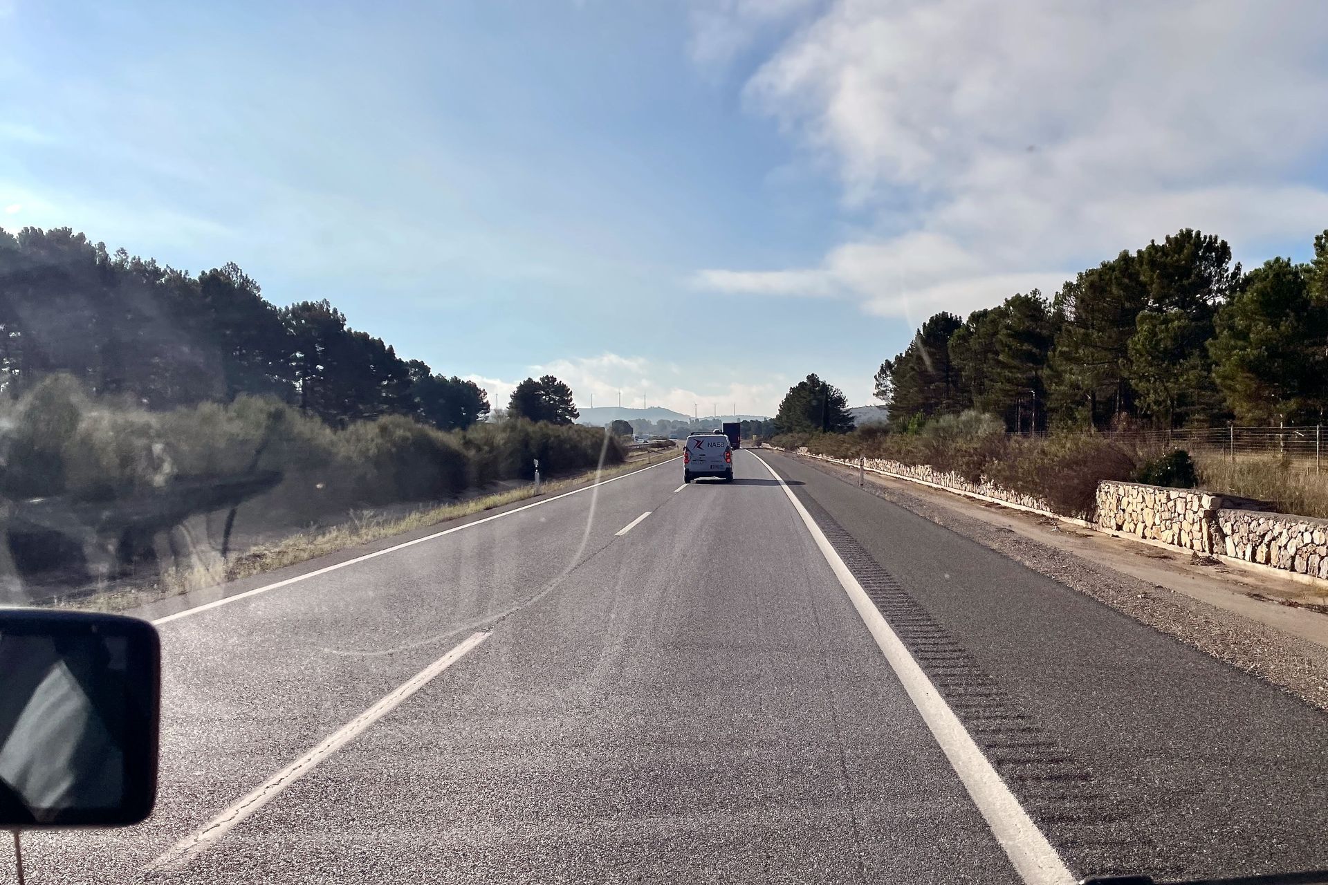 Wegbeeld met een voertuig dat voor je rijdt op een zonnige dag. Bomen langs de kant, de lucht is blauw.