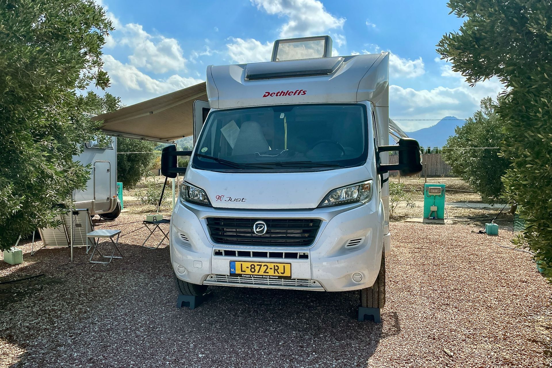 Ons plekje voor 4 nachtjes Witte camper geparkeerd op grind, luifel uitgeschoven, bomen en bergen op de achtergrond.