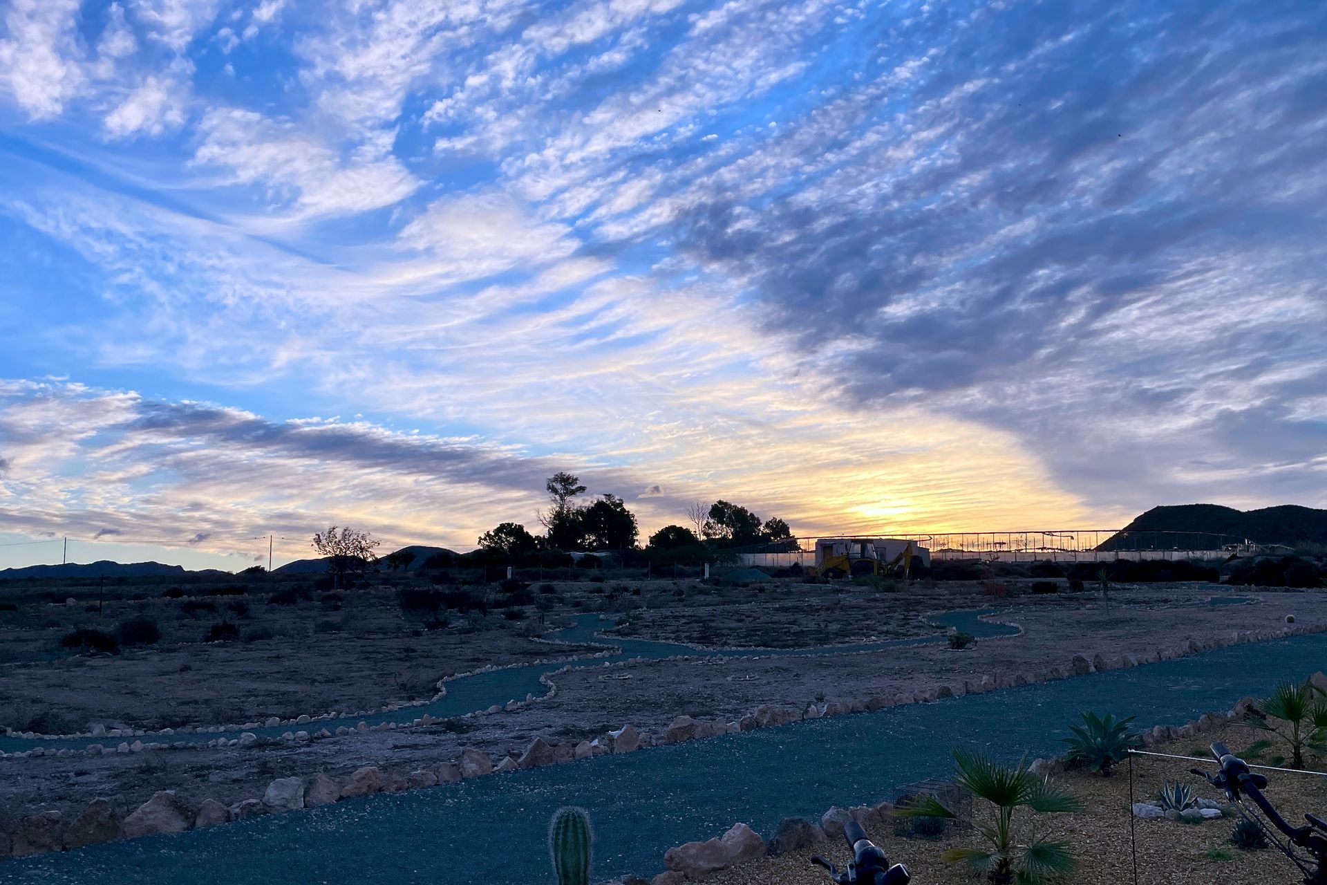 Zonsondergang over een droog landschap met strepenwolken en silhouetten van gebouwen en bomen.