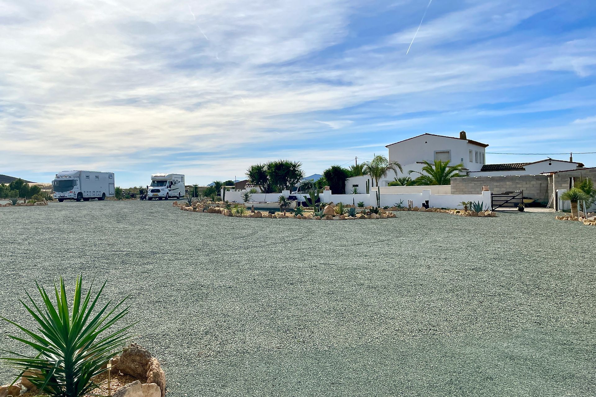 Grindparkeerplaats voor campers met twee campers, witte gebouwen en een blauwe lucht met wolken.