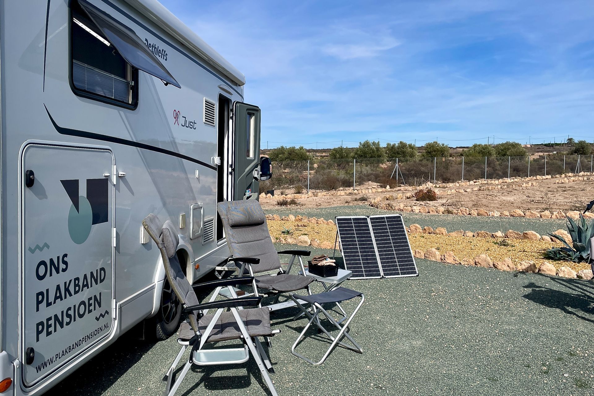 Camper geparkeerd op grind, met stoelen en zonnepanelen buiten. Zonnige dag.