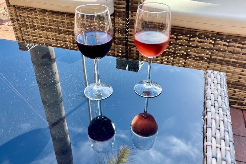 Twee wijnglazen, één rood en één rosé, op een spiegelende glazen tafel buiten.