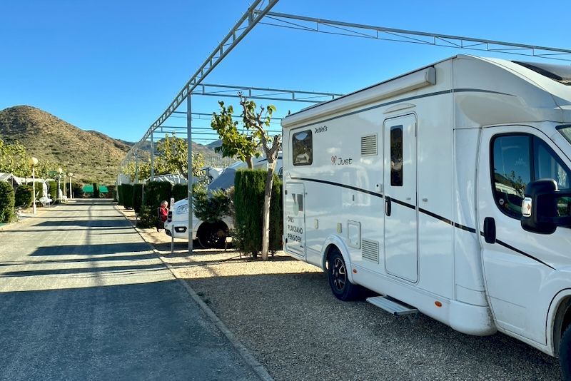 Witte camper geparkeerd op een grindterrein, onder een metalen luifel. Kampeerweg met groene heggen en bergen op de achtergrond.