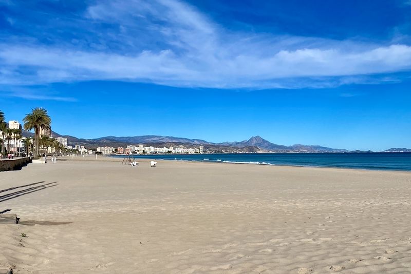 Een leeg strand met licht zand, helderblauw water en gebouwen langs de kust onder een blauwe lucht.