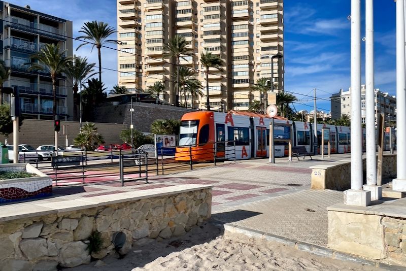 Oranje en witte tram bij een station aan de kust met palmbomen en een hoog gebouw op de achtergrond.