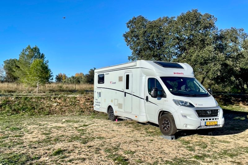 Witte camperbus geparkeerd op een onverhard terrein met bomen onder een blauwe lucht.