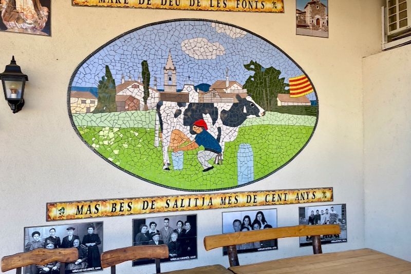 Mozaïek van een koe die gemolken wordt, met op de achtergrond een dorpstafereel, op een muur met borden en foto's.