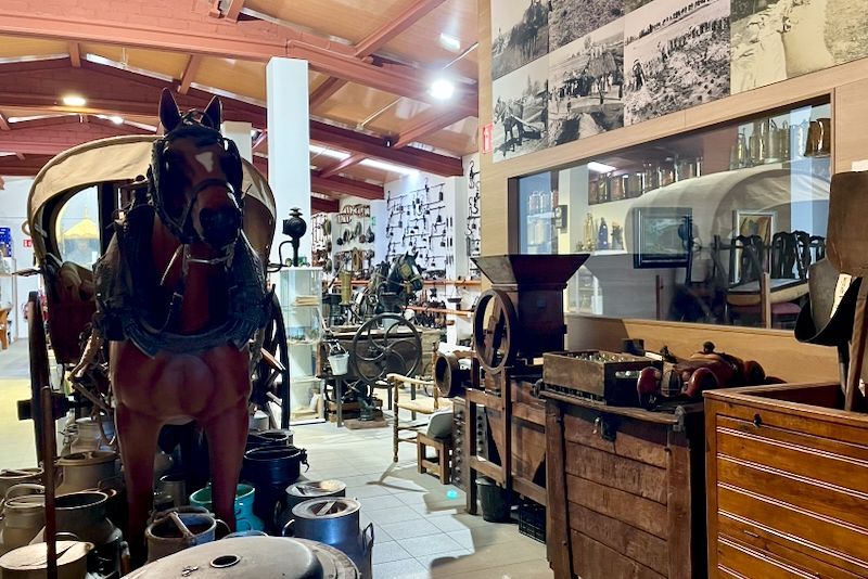 Museuminterieur met een tentoonstelling van door paarden getrokken rijtuigen, antieke gereedschappen en historische foto's.
