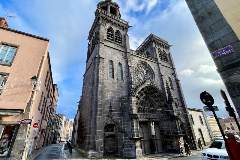 Buitenaanzicht van een kathedraal met twee hoge torens. Het gebouw is van grijze steen met sierlijke details. Bewolkte blauwe lucht.