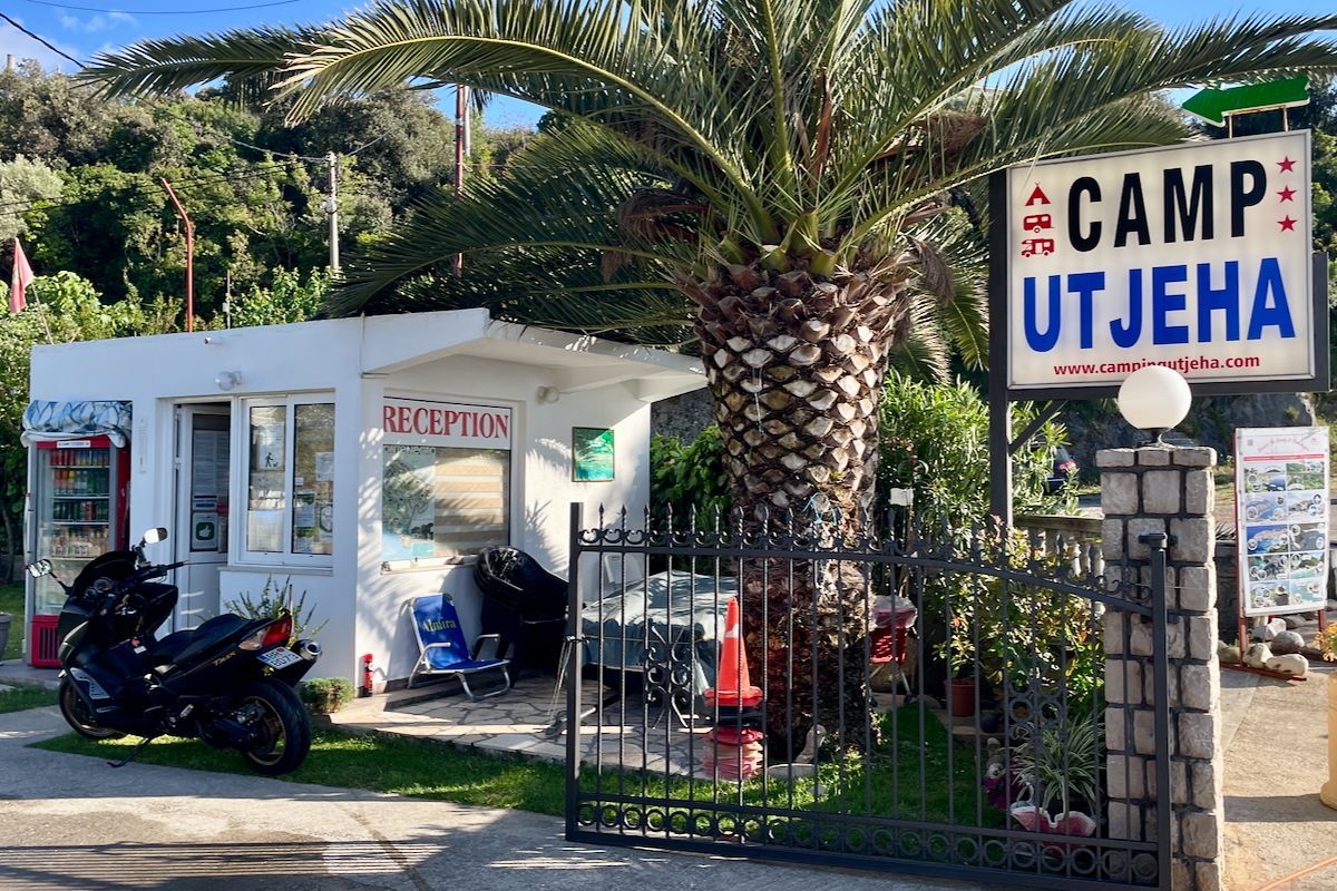 De ingang van de camping Een motorfiets staat geparkeerd voor een gebouw met een bord waarop staat: camp utjeha.
