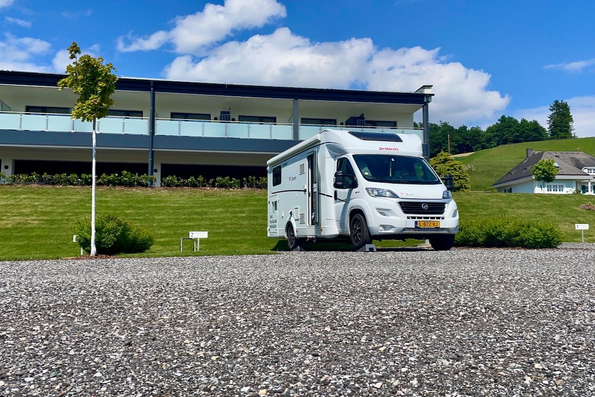 Onze overnachtingsplek Een witte camper staat geparkeerd voor een gebouw op een grindweg.