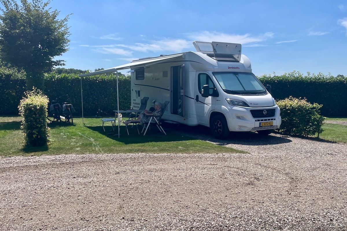 Onze plek voorlopig Een witte camper met luifel staat geparkeerd op een grindterrein.