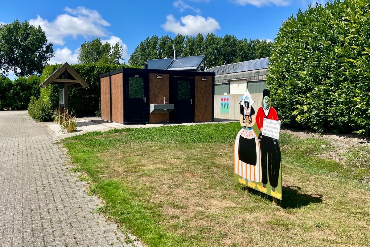 Een klederdracht fotopaneel staat op het gras voor een toiletgebouw.