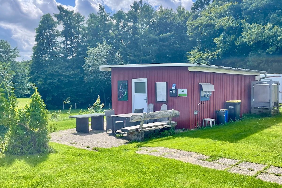 Informatie ruimte Een kleine, rode hut met een witte deur, bank en tafel staat in een grasveld. Bomen op de achtergrond op een zonnige dag.