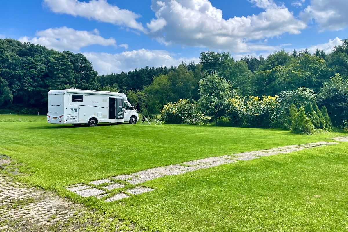 We zijn nu wel alleen Een witte camper geparkeerd op een grasveld met een pad. Bomen en een bewolkte lucht op de achtergrond.