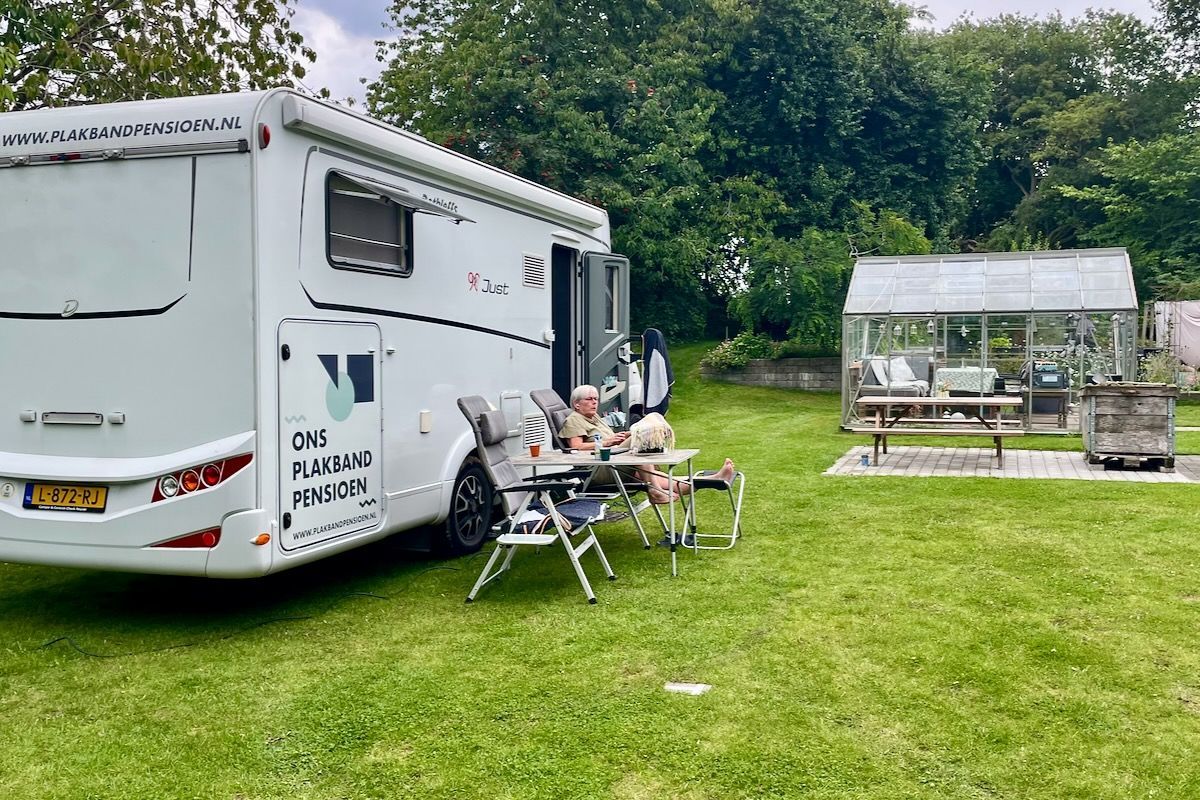 Ons plekje voor een week Een witte camper staat op het gras geparkeerd, met twee mensen die buiten aan een tafel zitten. Er staat een kleine kas in de buurt.