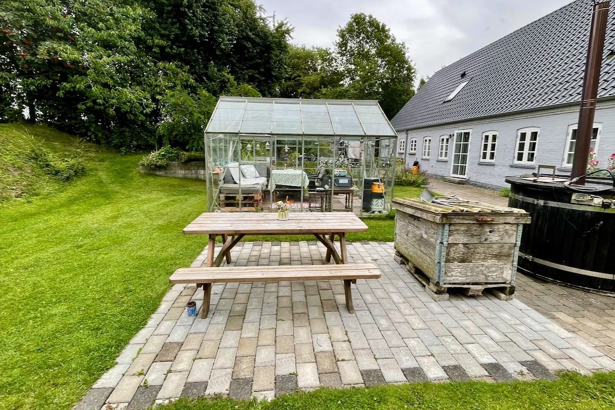 Terras en glazenhuis Een rustiek buitentafereel met een houten picknicktafel op een stenen terras, een kas en een boerderij.