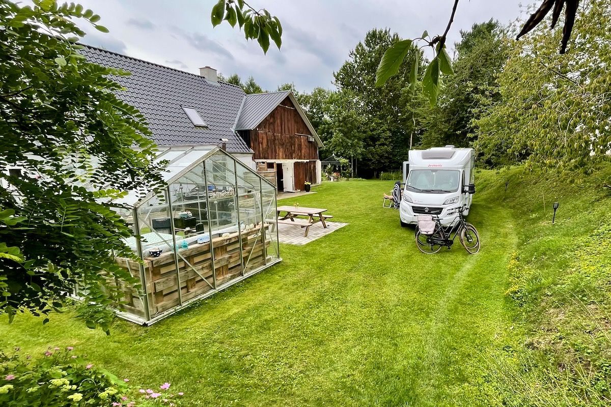 Uit de hoogte Een huis, een kas en een camper staan op een met gras begroeide heuvel. Erboven hangt een bewolkte lucht.