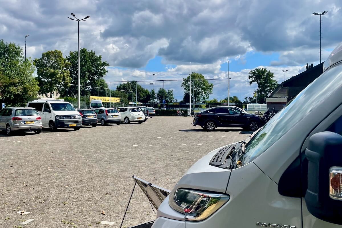 Cp met voetbalveld Parkeerterrein met diverse auto's op een grindpad, onder een gedeeltelijk bewolkte hemel.