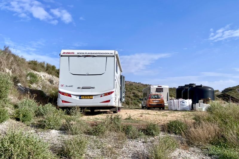Een camper staat op een heuvel geparkeerd met twee andere voertuigen, watertanks en bouwmaterialen onder een blauwe lucht.