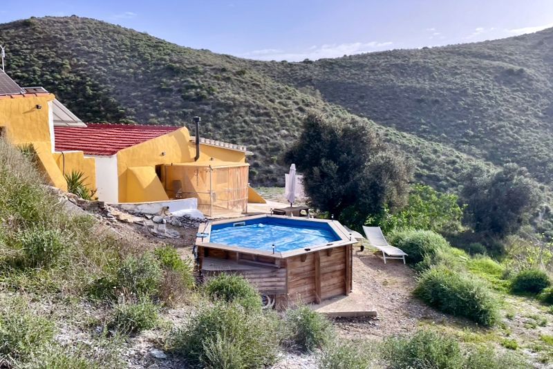 Geel huis met rood dak, houten jacuzzi, twee ligstoelen en uitzicht op de bergen.